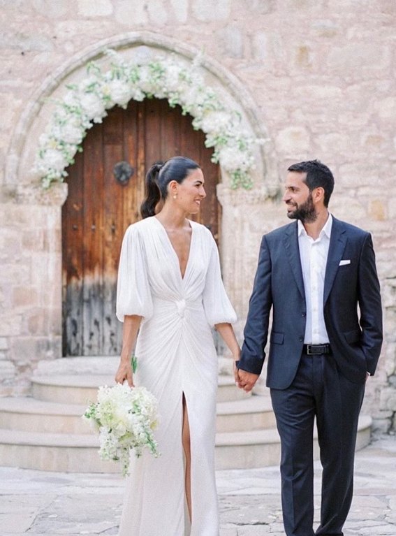 Bride and groom hand in hand in front of a stone chapel with a white floral arch in Cyprus